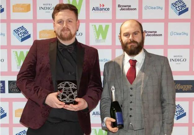 Two men are standing in front of a pink backdrop with various logos; one man on the left in a maroon patterned suit is holding a dark award plaque, and the man on the right in a grey three-piece suit is holding a bottle of champagne. This image represents the 'Winner' recognition for 'Business Services' at the Exeter Living Awards 2024.