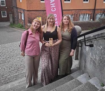 Three women standing on stone steps outdoors, one in the middle holding a wooden award plaque, with a pink banner in the background. This image represents the 'Highly Commended' recognition for 'Business of the Year' at the Exeter Chamber Impact Awards 2025.