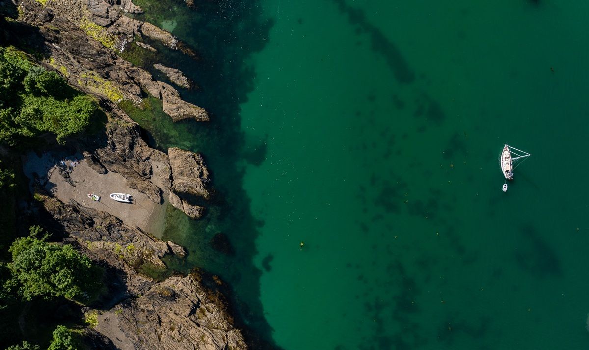 An aerial photo of a cove in Cornwall with 2 boats on the beach and a sail boat moored up.  A person is swimming.