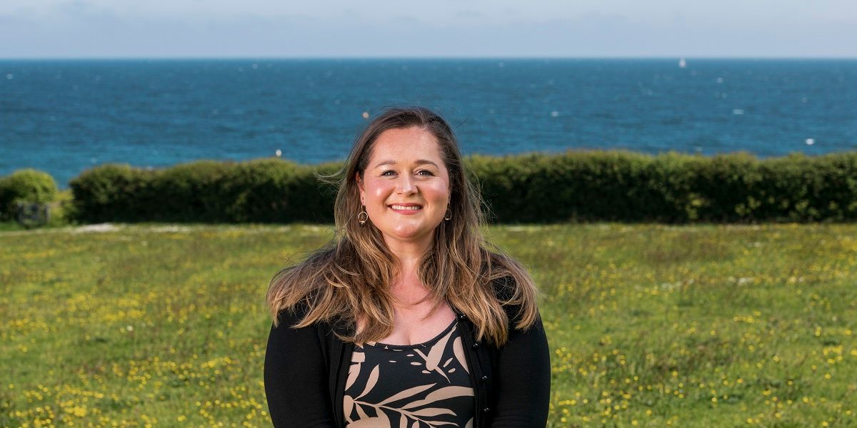 Freya Randall-Money stands in a field with hedge and then view out to sea, Falmouth Bay Cornwall