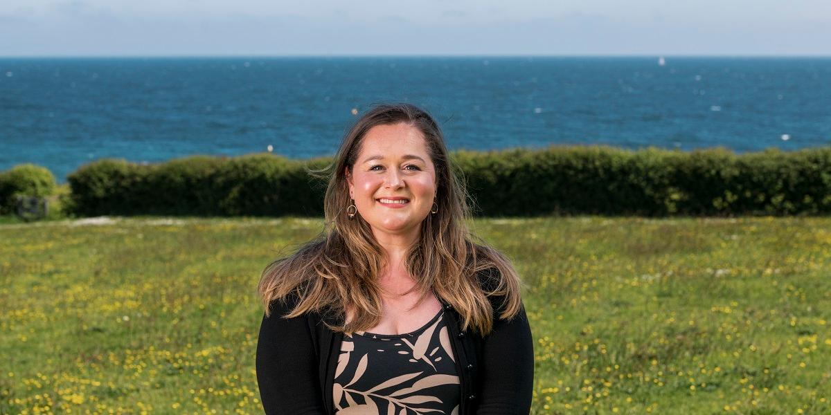 Freya Randall-Money stands in a field with hedge and then view out to sea, Falmouth Bay Cornwall