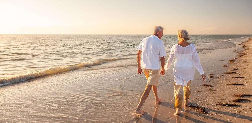 A man and woman in their 60s walk along a beach holding hands as the sun sets