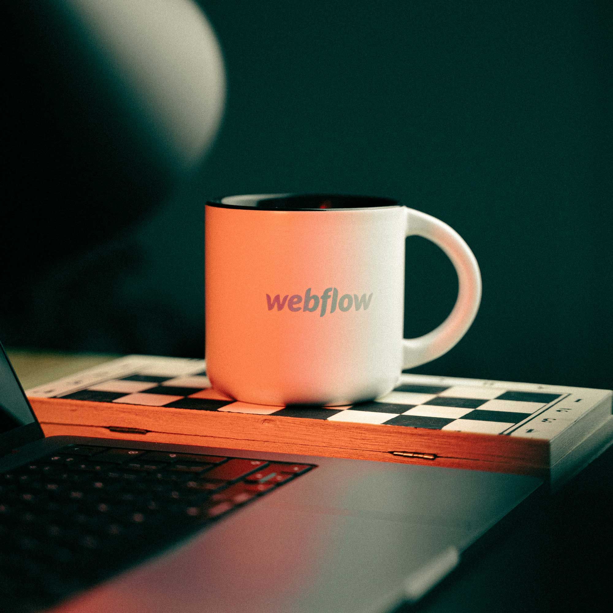 A white Webflow mug with a colorful logo sits on a chessboard next to a laptop.