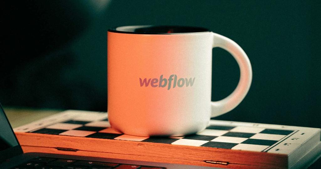 A white Webflow mug with a colorful logo sits on a chessboard next to a laptop.