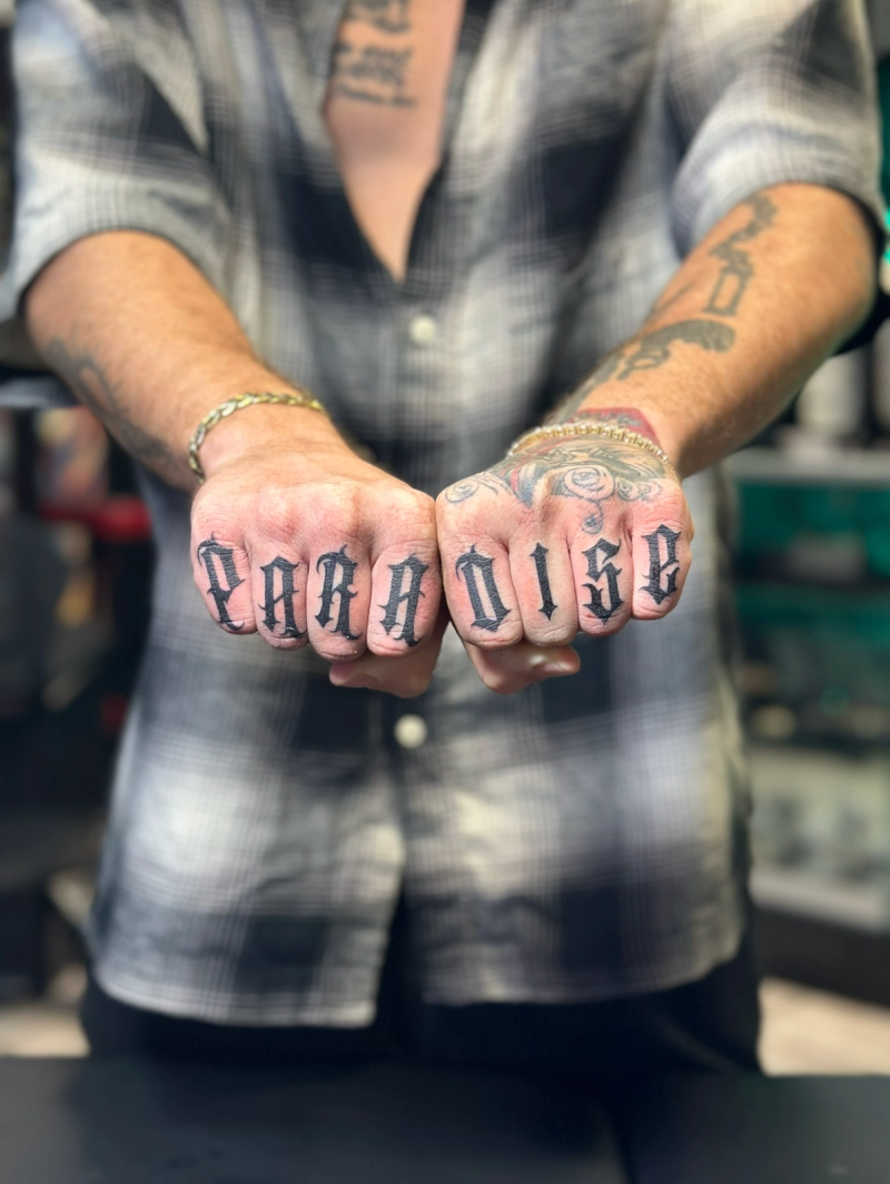 Black and grey script lettering tattoo on knuckles spelling 'paradise'