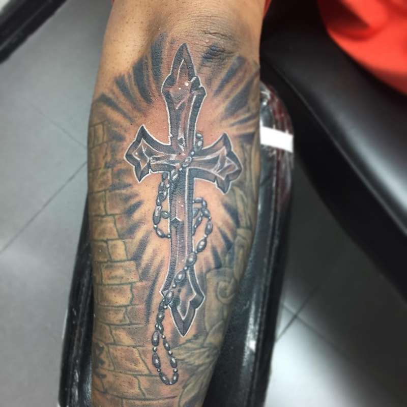 Black and grey illustrative cross tattoo with rosary on forearm
