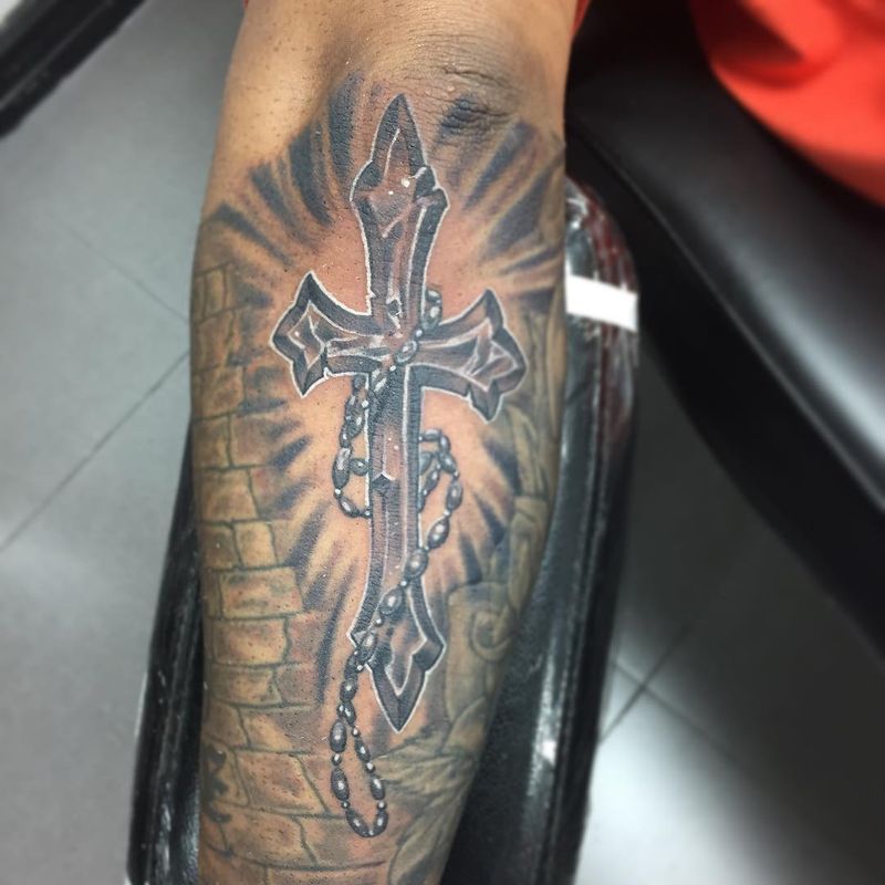 Black and grey illustrative cross tattoo with rosary on forearm