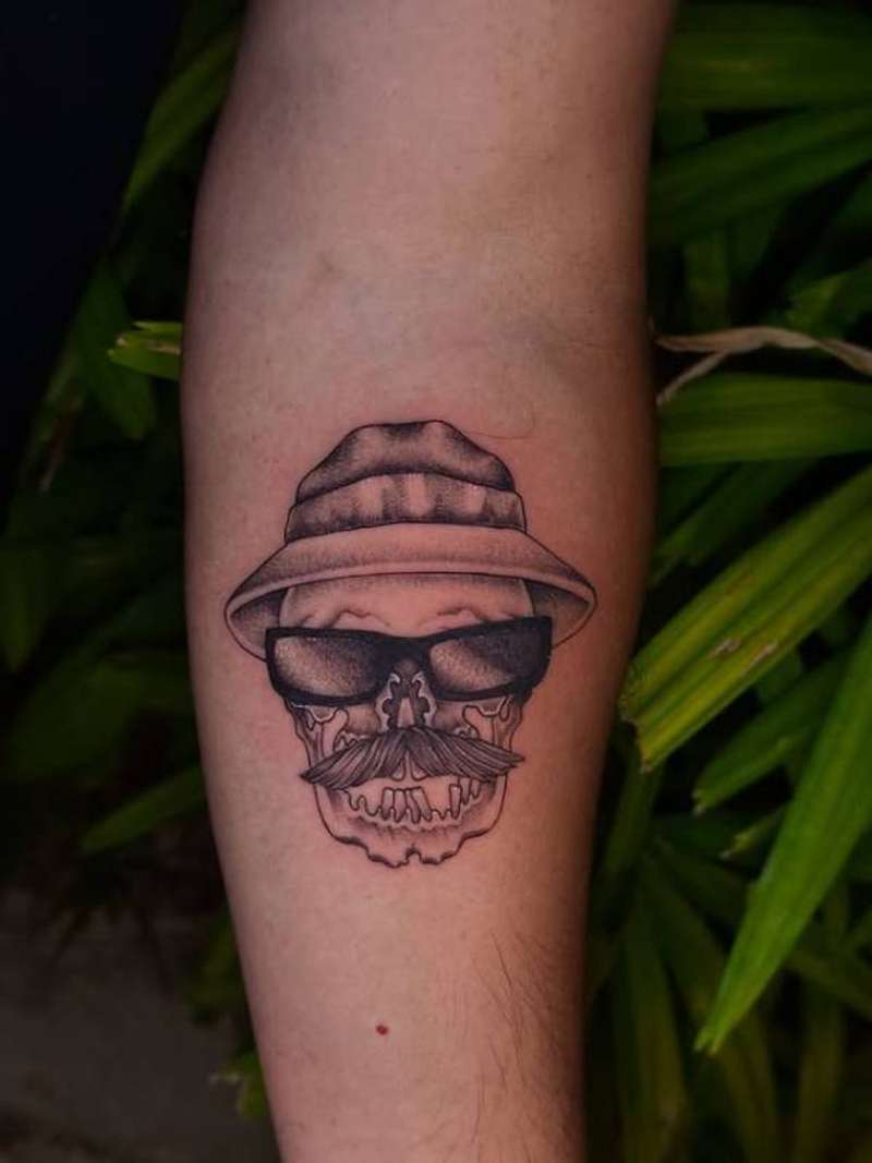 Black and grey illustrative skull with hat and sunglasses tattoo on forearm