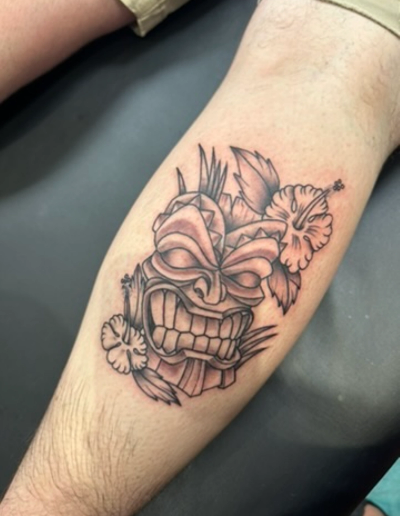 Black and grey illustrative tiki mask and floral tattoo on calf