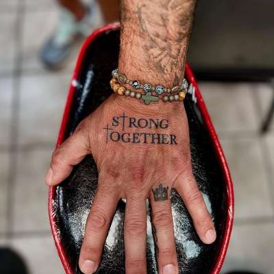 Black and grey lettering tattoo on hand reading 'Strong Together'