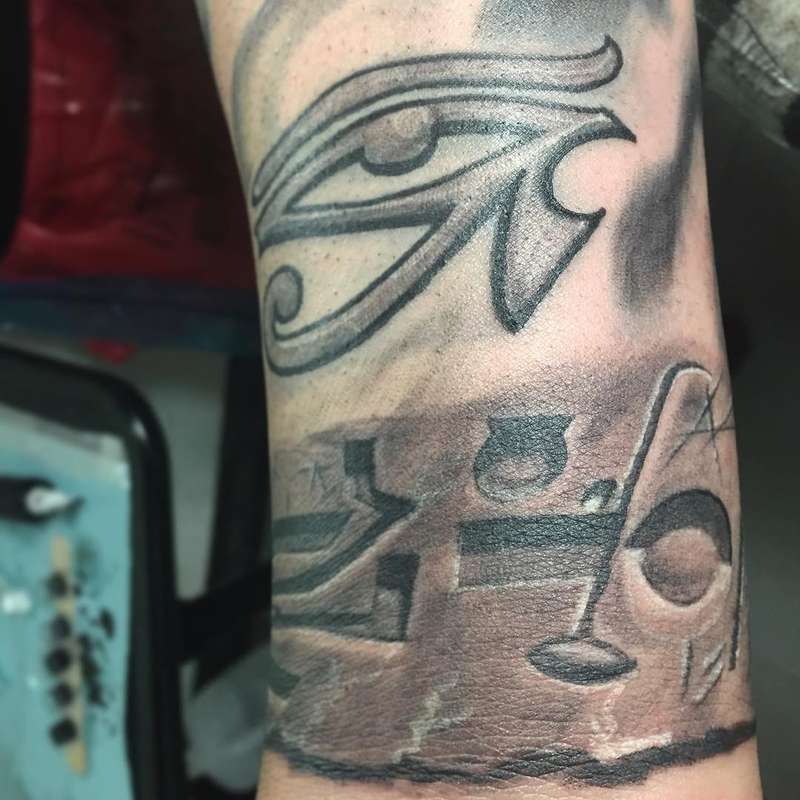 Black and grey Eye of Horus tattoo with hieroglyphs on forearm.