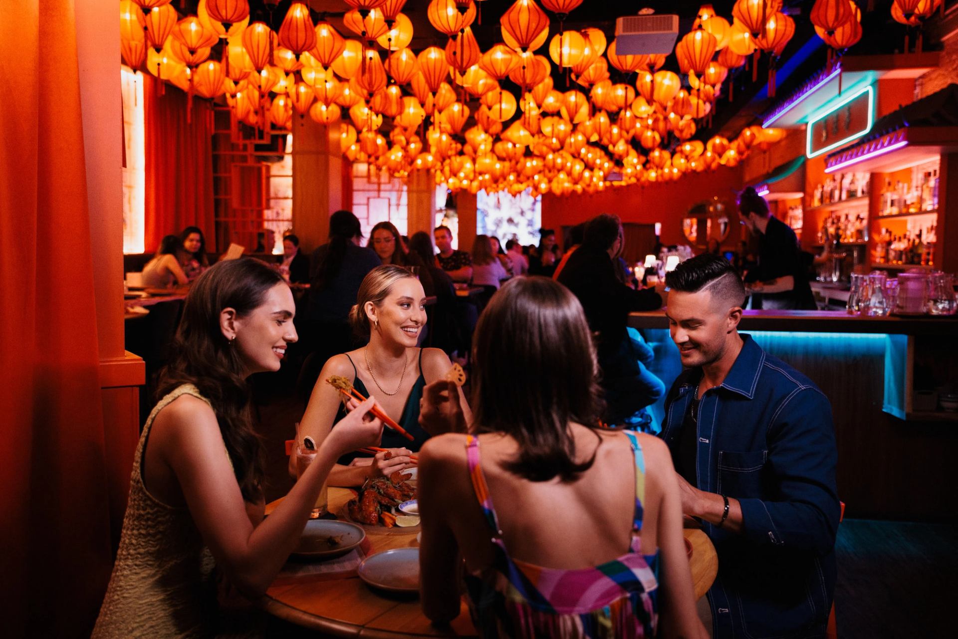 Four friends dine at an upscale restaurant under the glow of orange lanterns hanging from the ceiling.