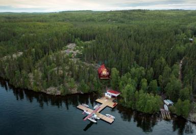 Aerial view of a small boat dock on the shore of a lake, with a lodge nearby surrounded in boreal forest.