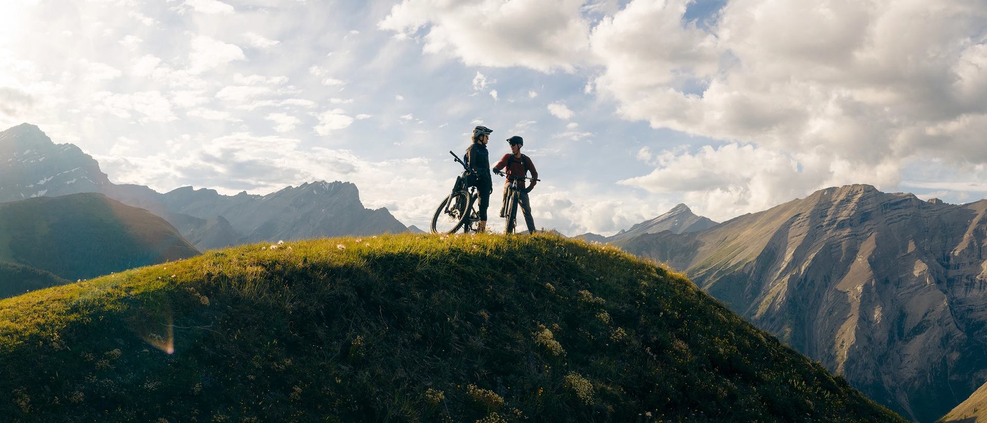 Two mountain bikers stand with their bikes on a grassy ridge at Fortress Mountain, silhouetted against a bright sky with scattered clouds and rugged peaks in the background.