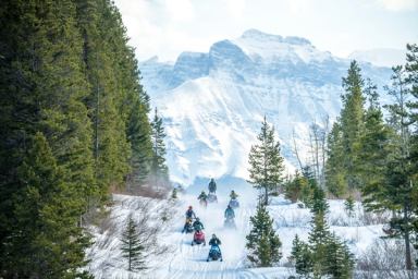 Snowmobilers ride through a snowy forest with majestic mountains in the background.