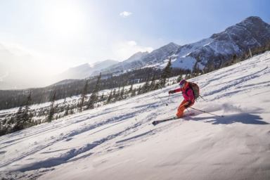 Two cyclists catch air while fat-biking across a snowy landscape with the Rocky Mountains behind them.