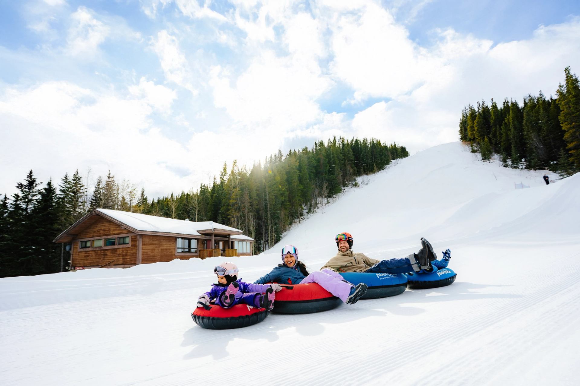 A family laughs and holds on while tubing side-by-side down a snowy hill at Nakiska.