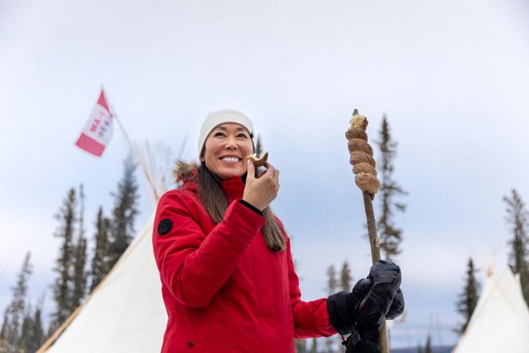 A women eating Bannock bread in front of a tipi during winter at the Aurora Borealis Indigenous Village.