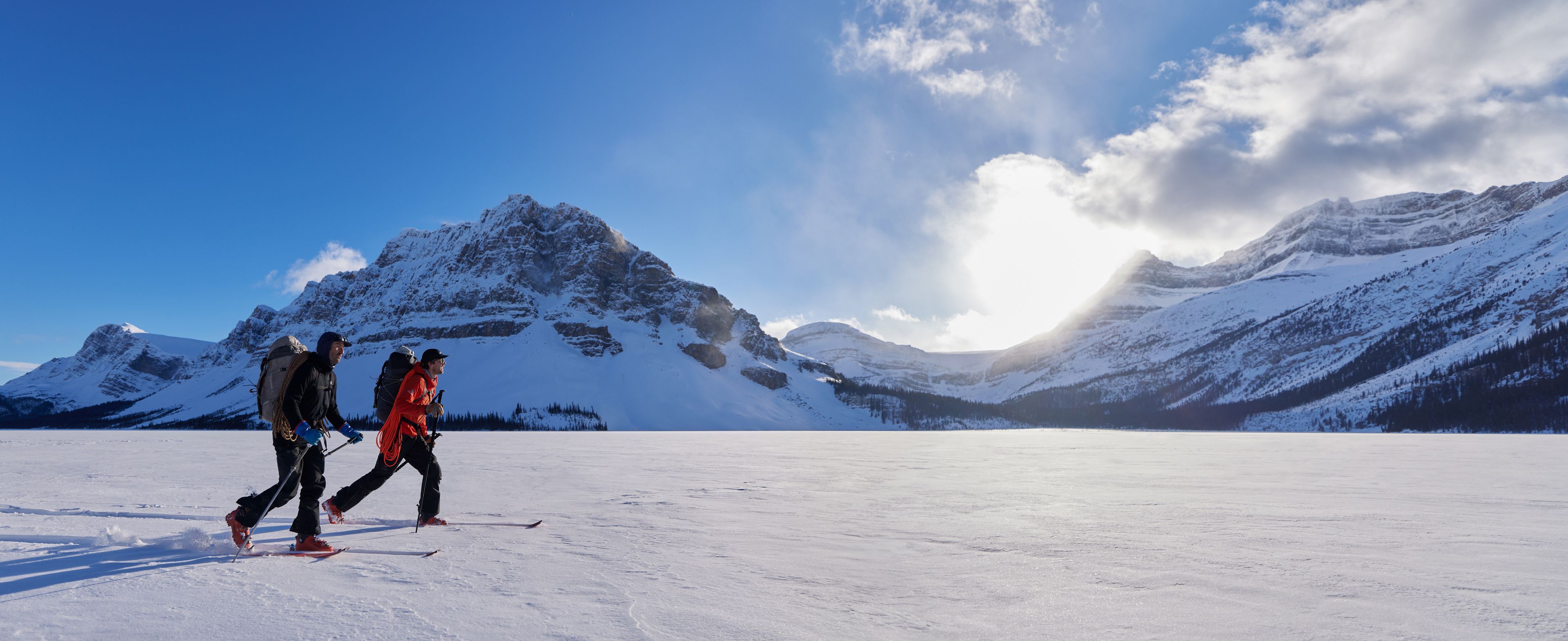 Two people cross-country ski across a wide snowy landscape with mountain peaks and bright sunlight in the background.