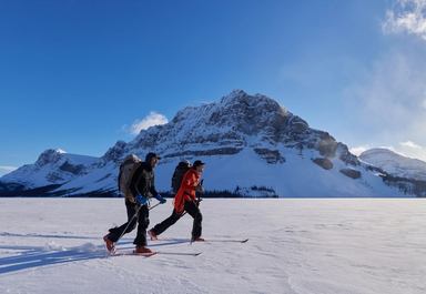 Two people cross-country ski across a wide snowy landscape with mountain peaks and bright sunlight in the background.