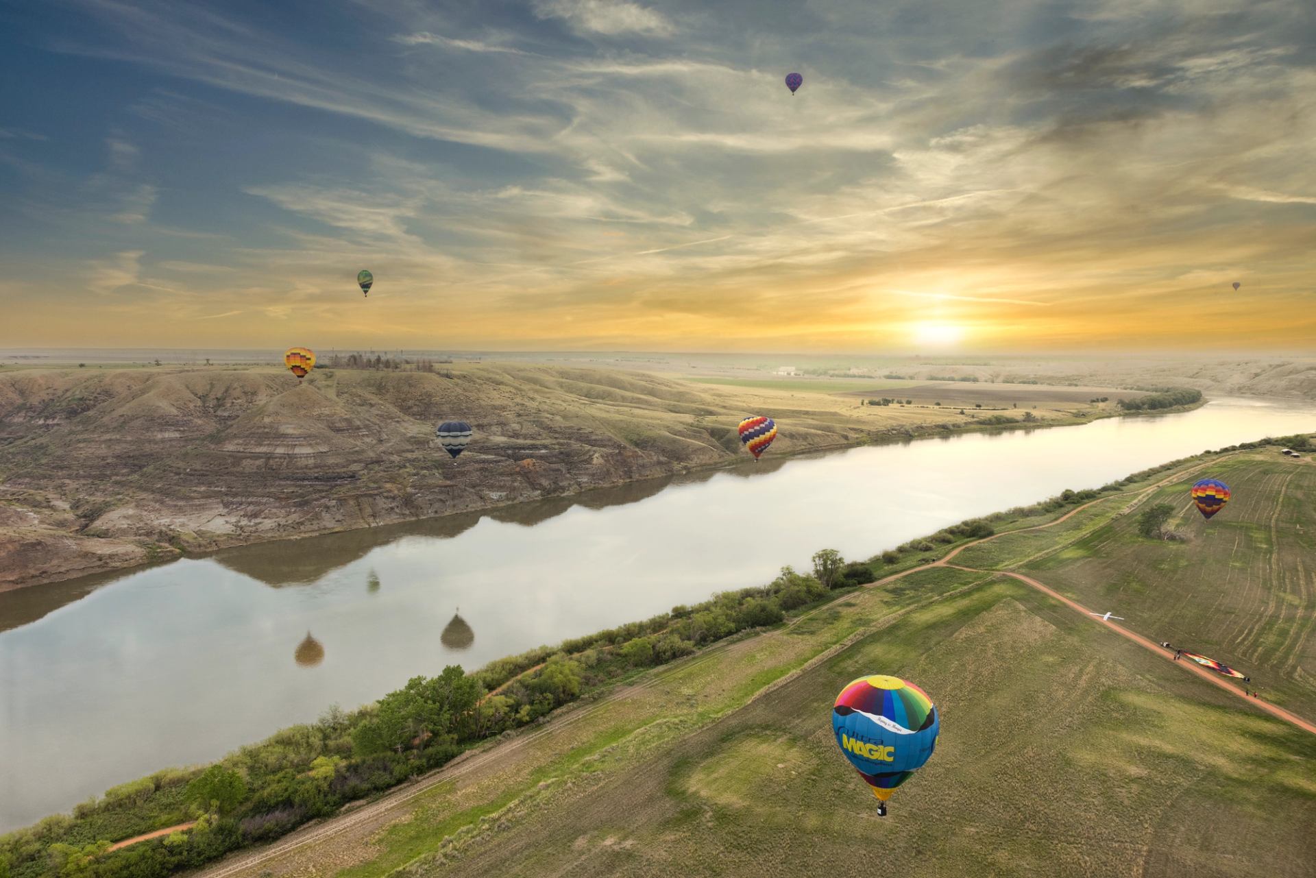 Aerial view of multiple colorful hot air balloons over a river with the sun setting.