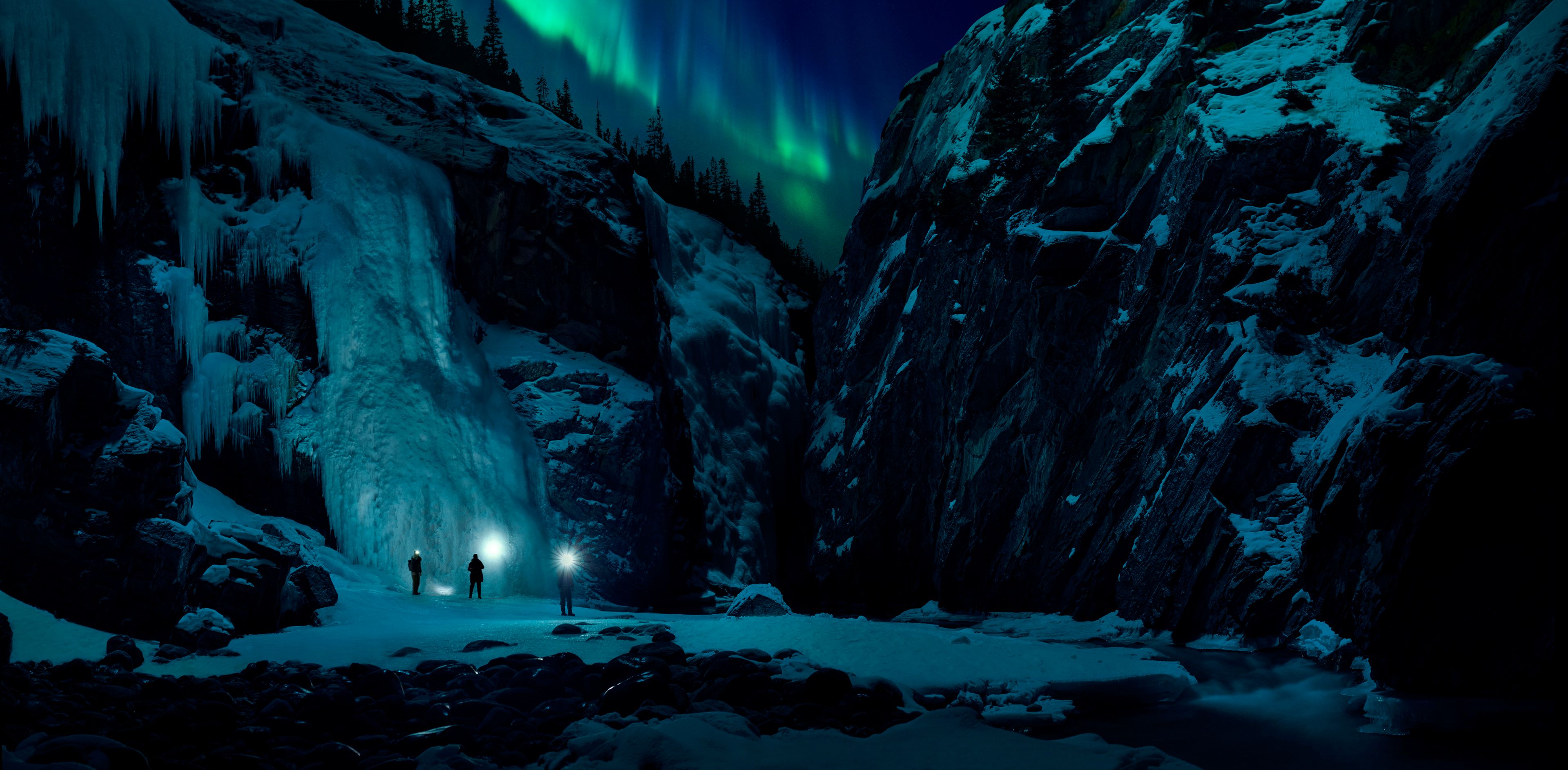 Group of three friends exploring a frozen canyon at nights with the Northern Lights overhead in David Thompson Country