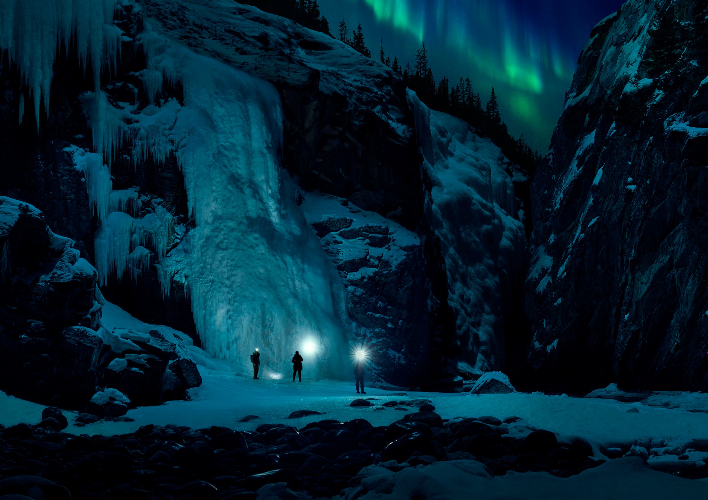 Group of three friends exploring a frozen canyon at nights with the Northern Lights overhead