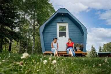 Guests sit outside a blue microcabin near a forested area.