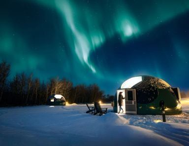 A woman stands in the doorway of a glamping dome looking up at the aurora borealis. 