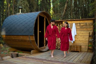 A couple walks out of a barrel sauna holding hands at the outdoor Wilderness Spa at The Woods Experience.