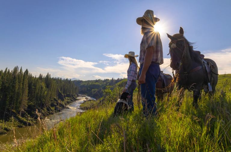 Two ranchers leading two horses and a dog overlook a riverbank.