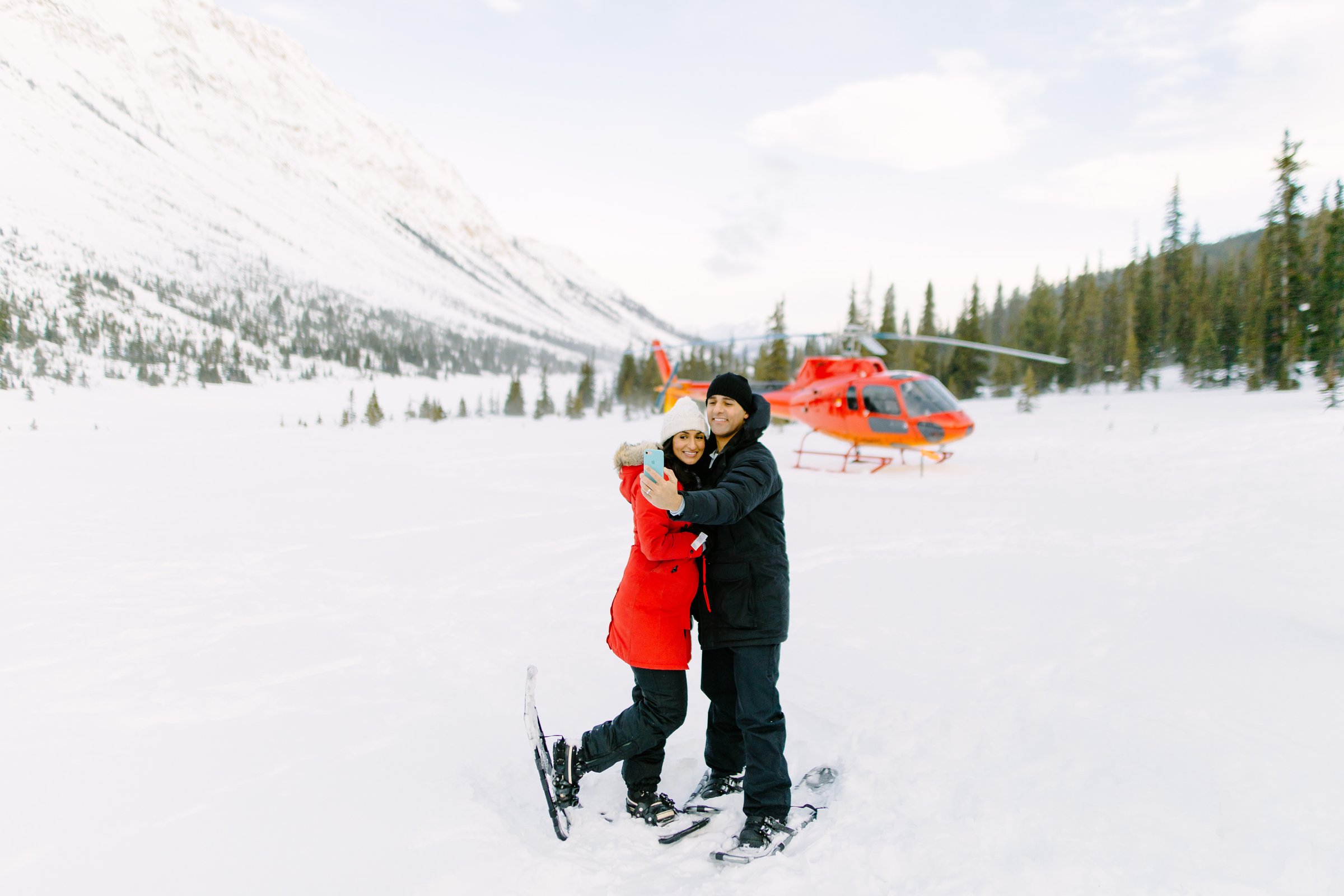 A couple take a selfie in front of an orange helicopter on a snowy mountain.
