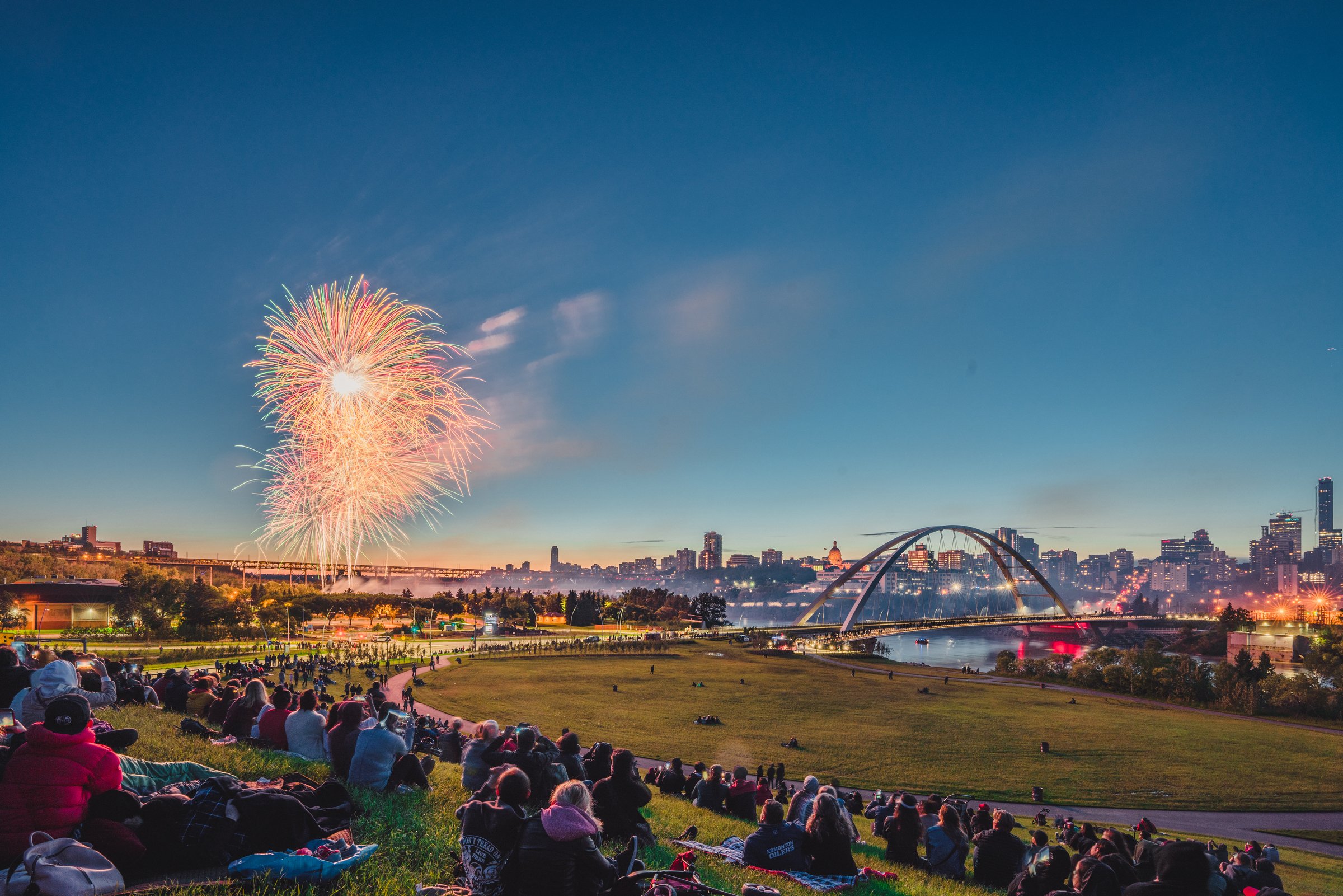 A crowd of people watching fireworks while sitting on the grass beside the North Saskatchewan River in Edmonton