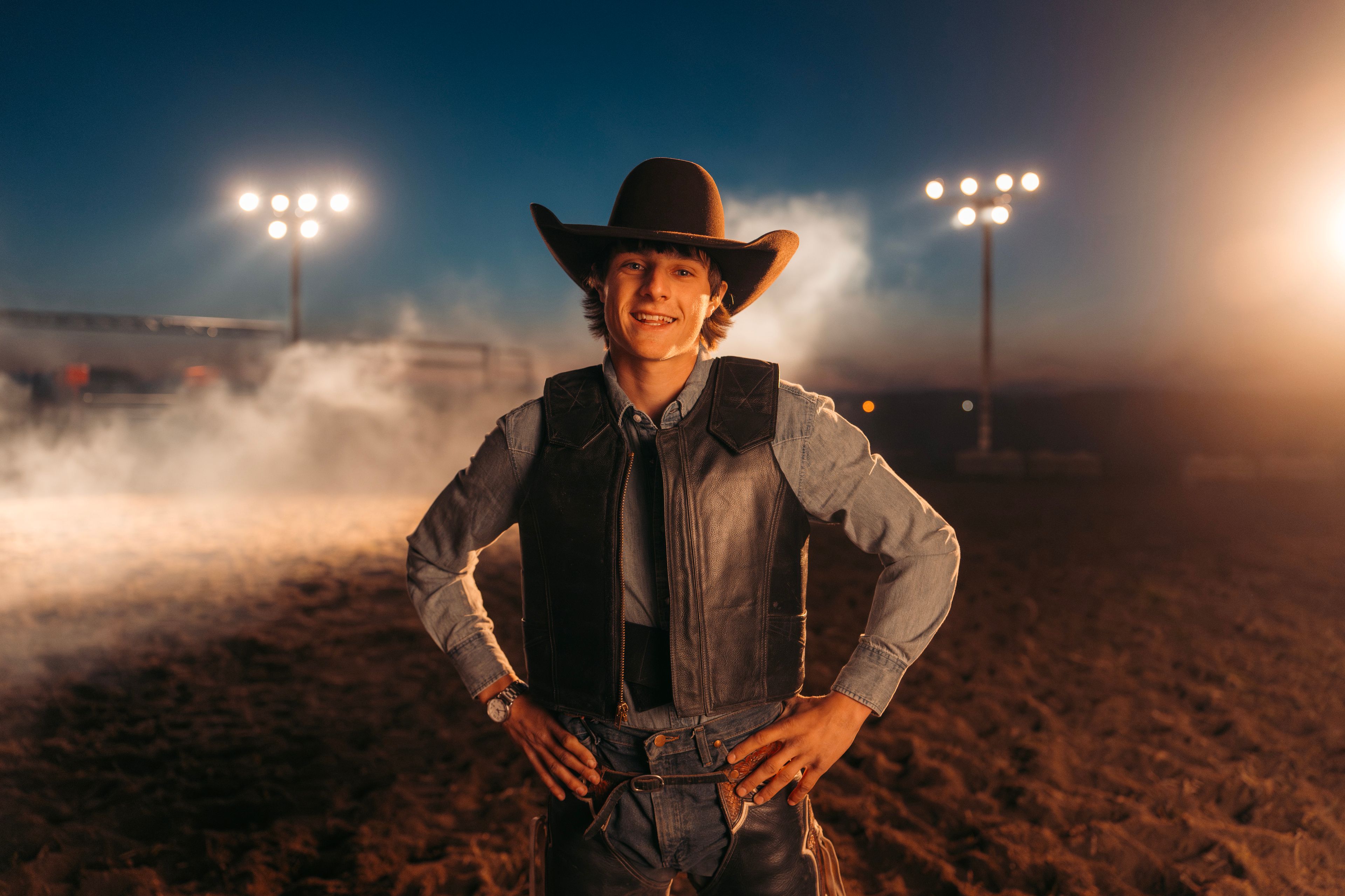 A bull rider stands with hands on hips in an arena at night.