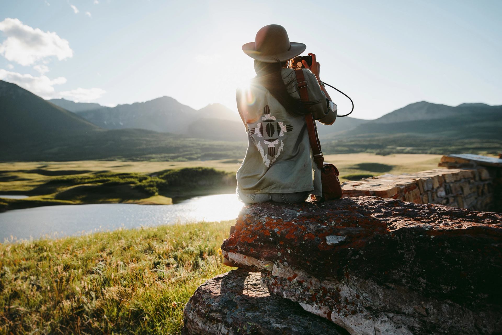 A woman sitting on a rock and taking a picture of a lake with mountains in the background