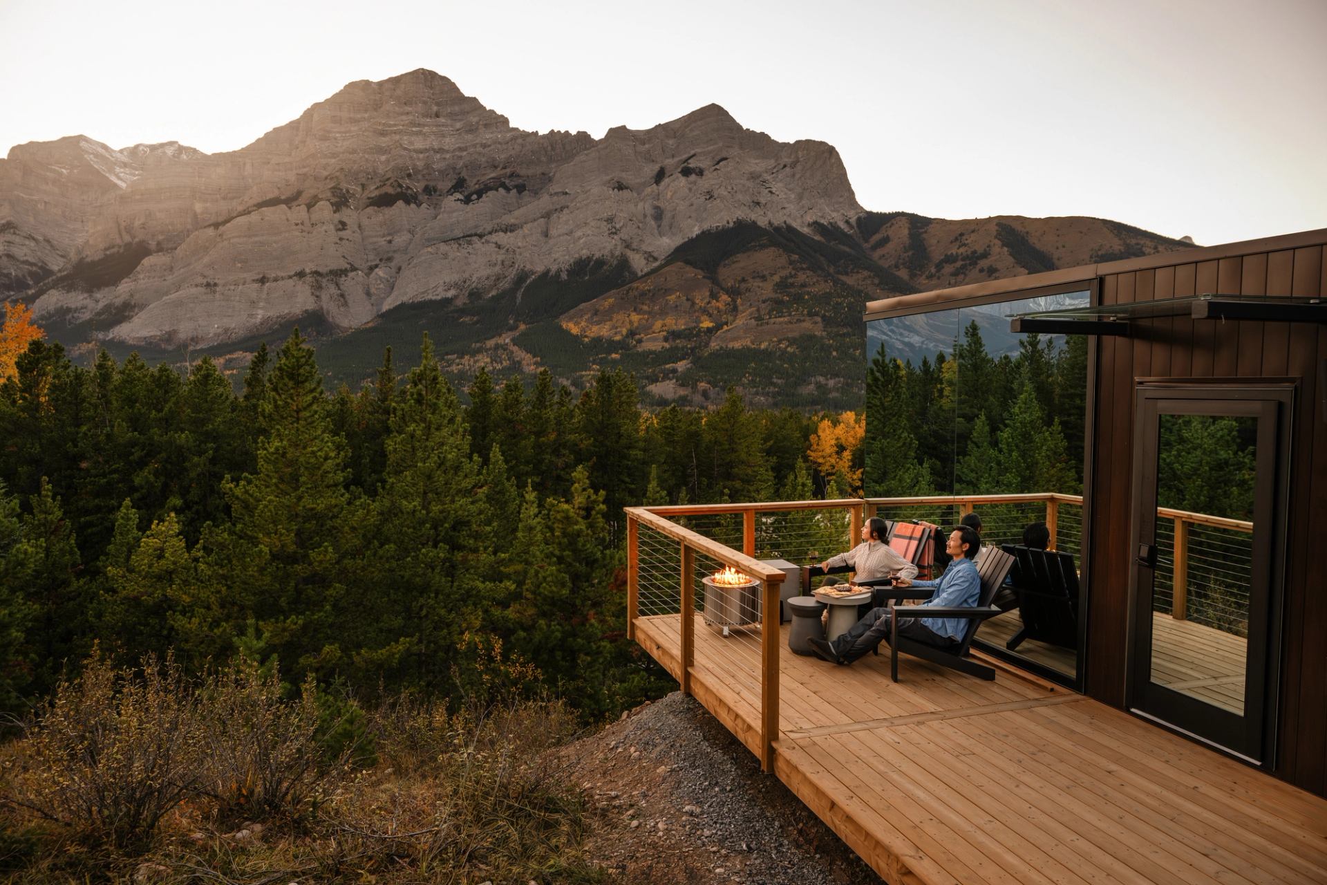 A couple sits on the deck at Skyridge Glamping in Kananaskis, with mountains, a forest, and sunset in the background.