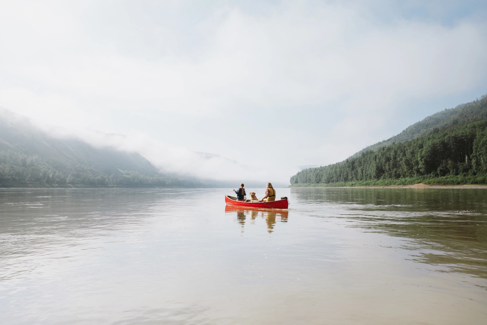 Couple / man and woman and dog paddling on a lake in a red canoe with misty forest and mountain scene in the background