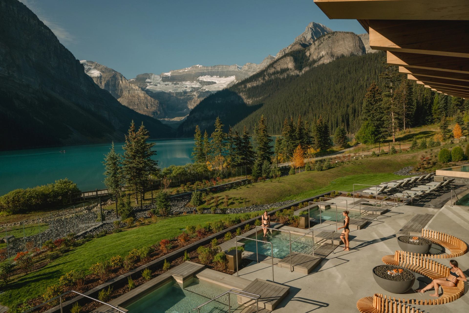 A wide shot of 2 people enjoying the outdoor pools at BASIN with Lake Louise in the background.