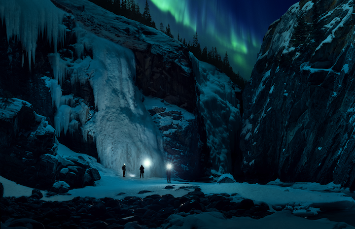 Group of three friends exploring a frozen canyon at nights with the Northern Lights overhead.