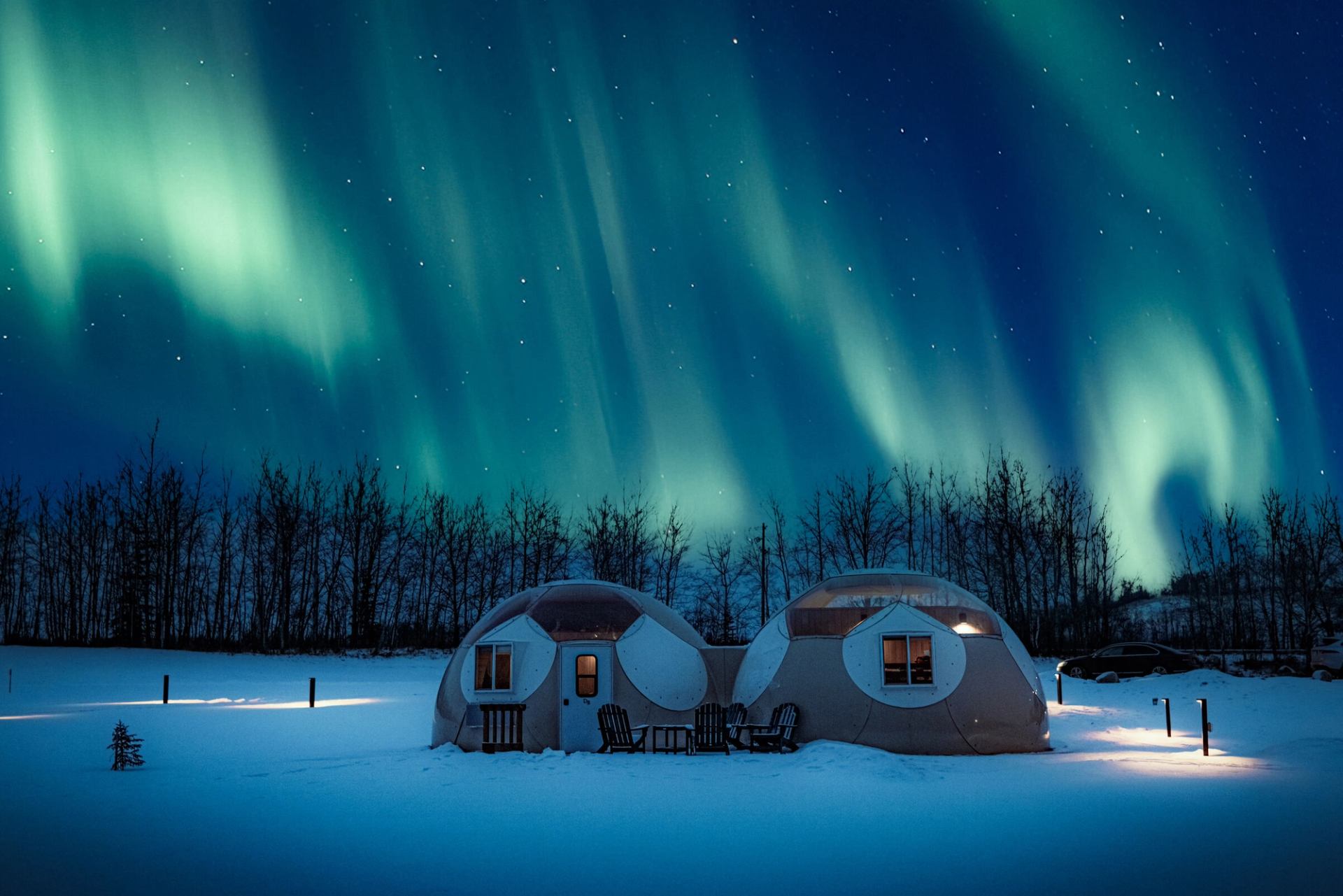Under the bright green streaks of the northern lights, two glamping domes are surrounded by snow.