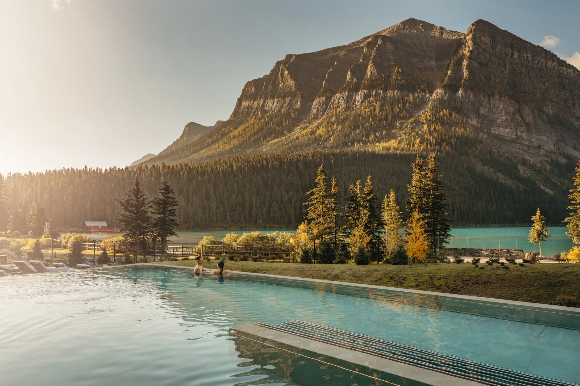 Two people stand in an outdoor pool overlooking Lake Louise and Fairview Mountain at sunset.