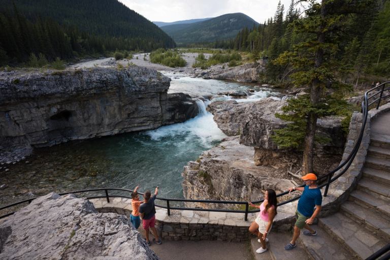 People stand on stone walkways beside rushing rapids, rocky cliffs and forested mountains.