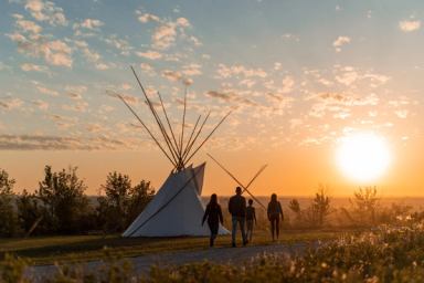 A family walks along a path toward a tipi at sunset in a natural outdoor setting.
