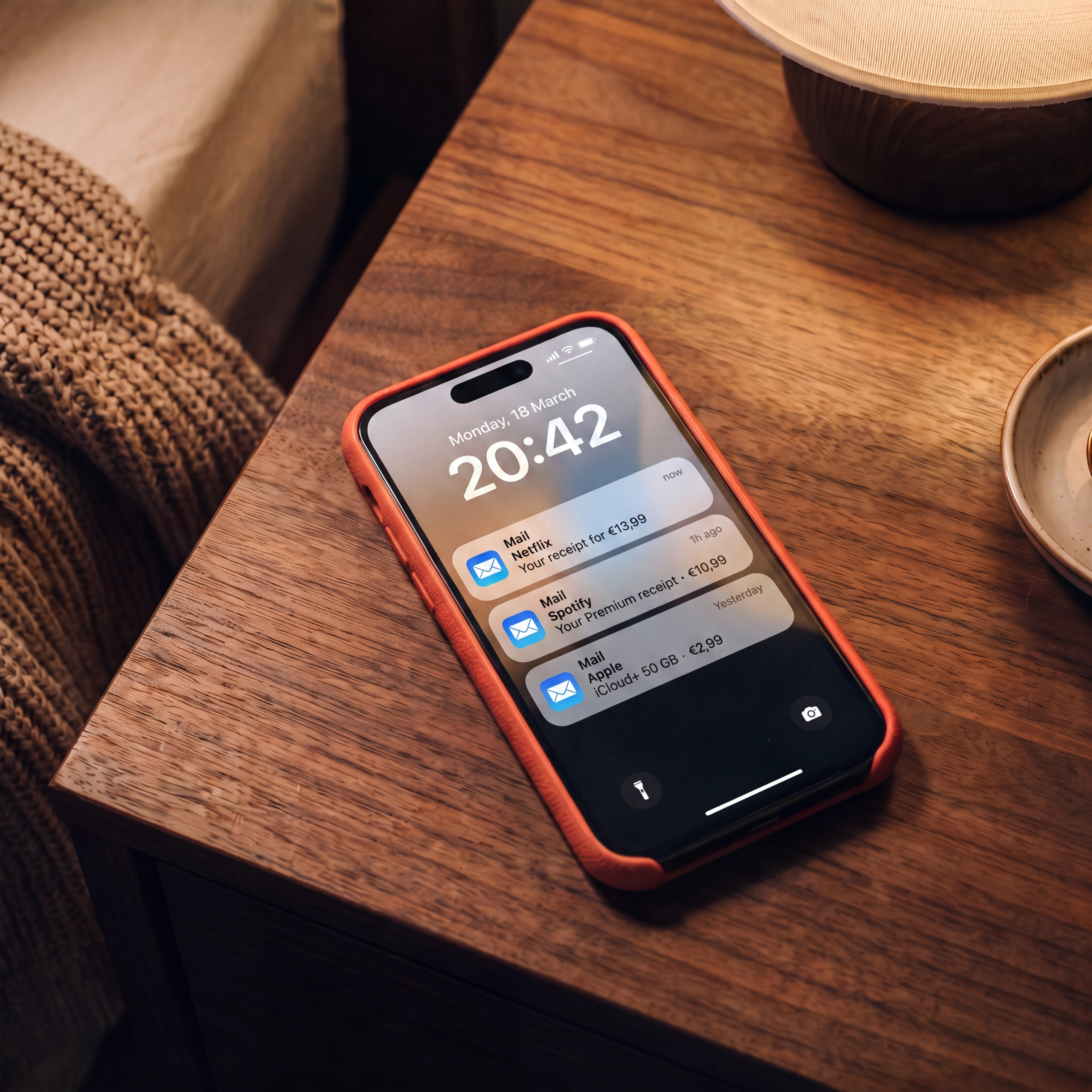 A coral iPhone on a wooden nightstand at night, lock screen showing Mail receipts for Netflix €13,99, Spotify €10,99 and iCloud+ €2,99.