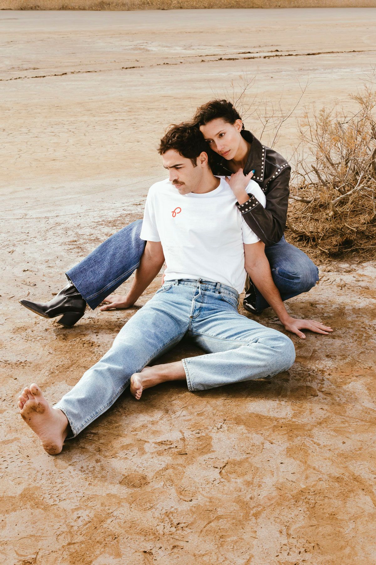 A couple sitting close on cracked, sunlit ground, one leaning back into the other.