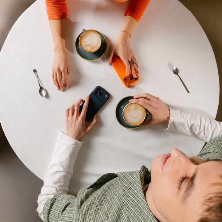 Seen from above, a couple at a small round café table: two coffees, two phones, four hands.
