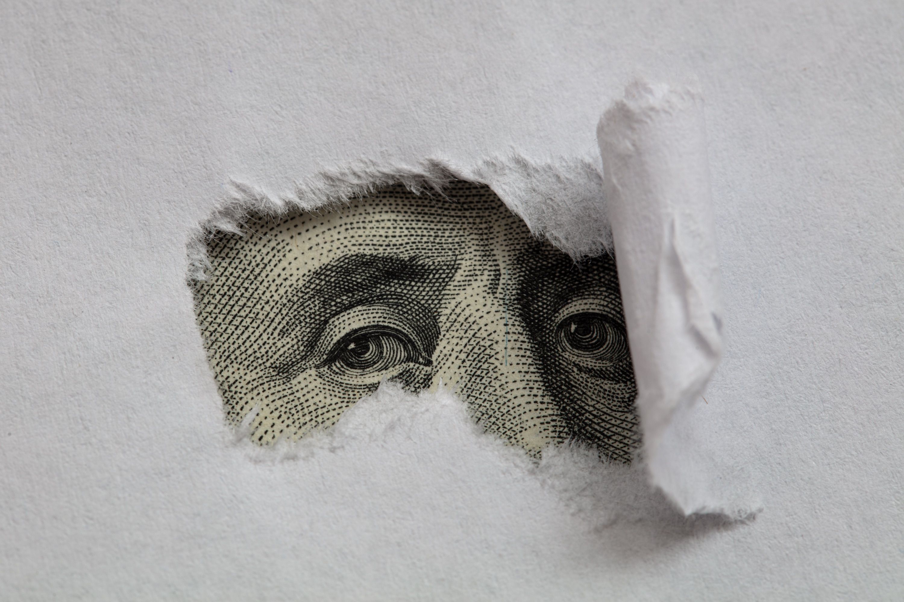 A dollar bill's engraved eyes looking out through a hole in torn white paper.