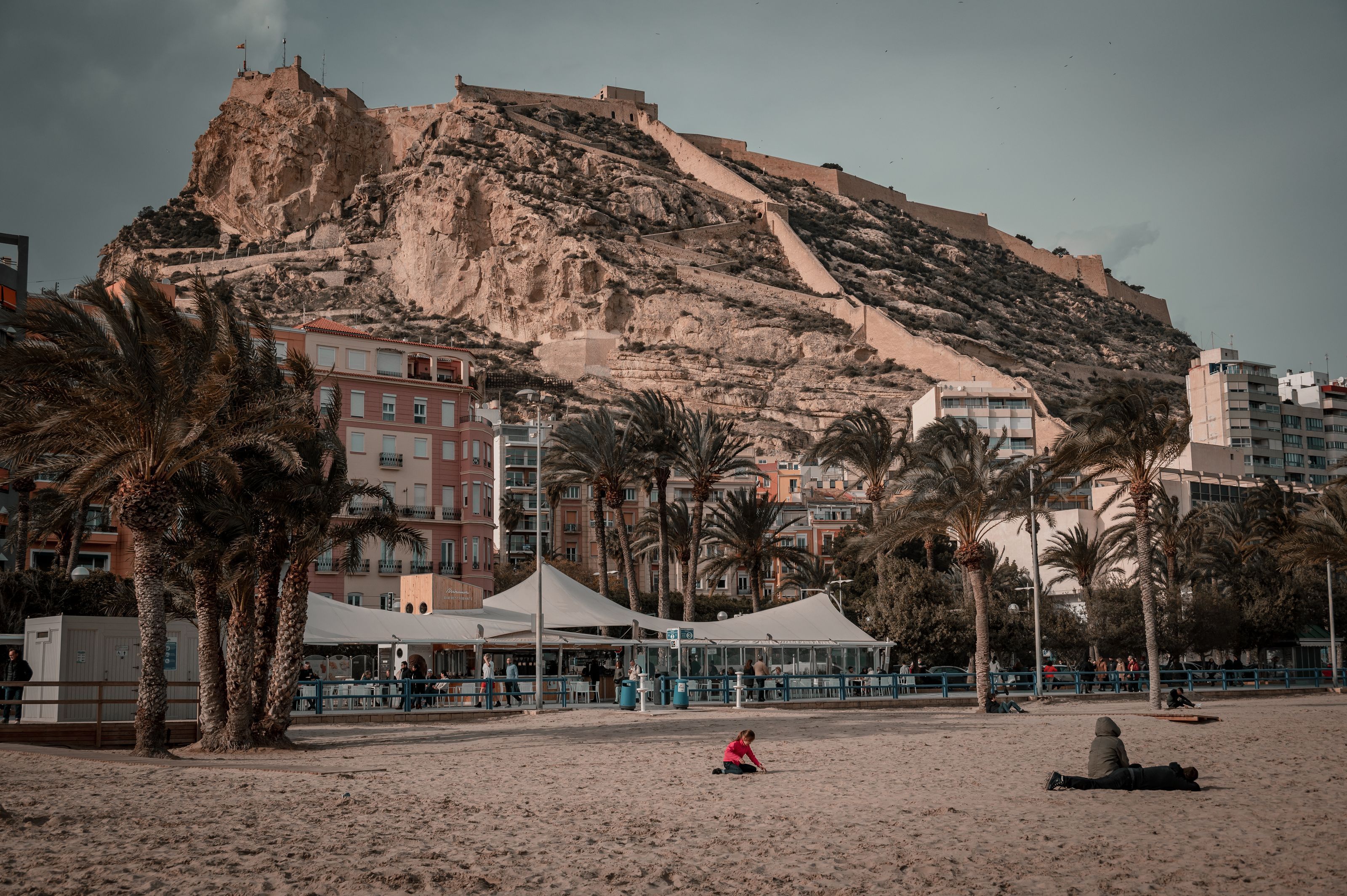Alicante's city beach on an overcast day, with palm trees, a castle on the hill above and a few people sitting on the sand in jackets