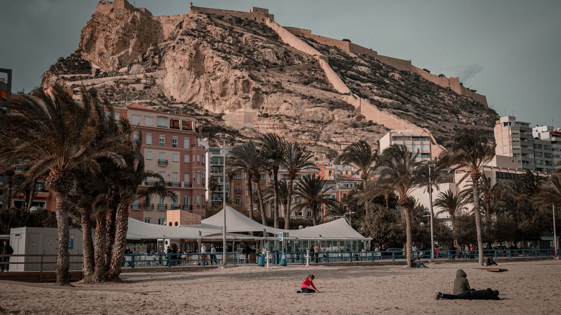 Alicante's city beach on an overcast day, with palm trees, a castle on the hill above and a few people sitting on the sand in jackets