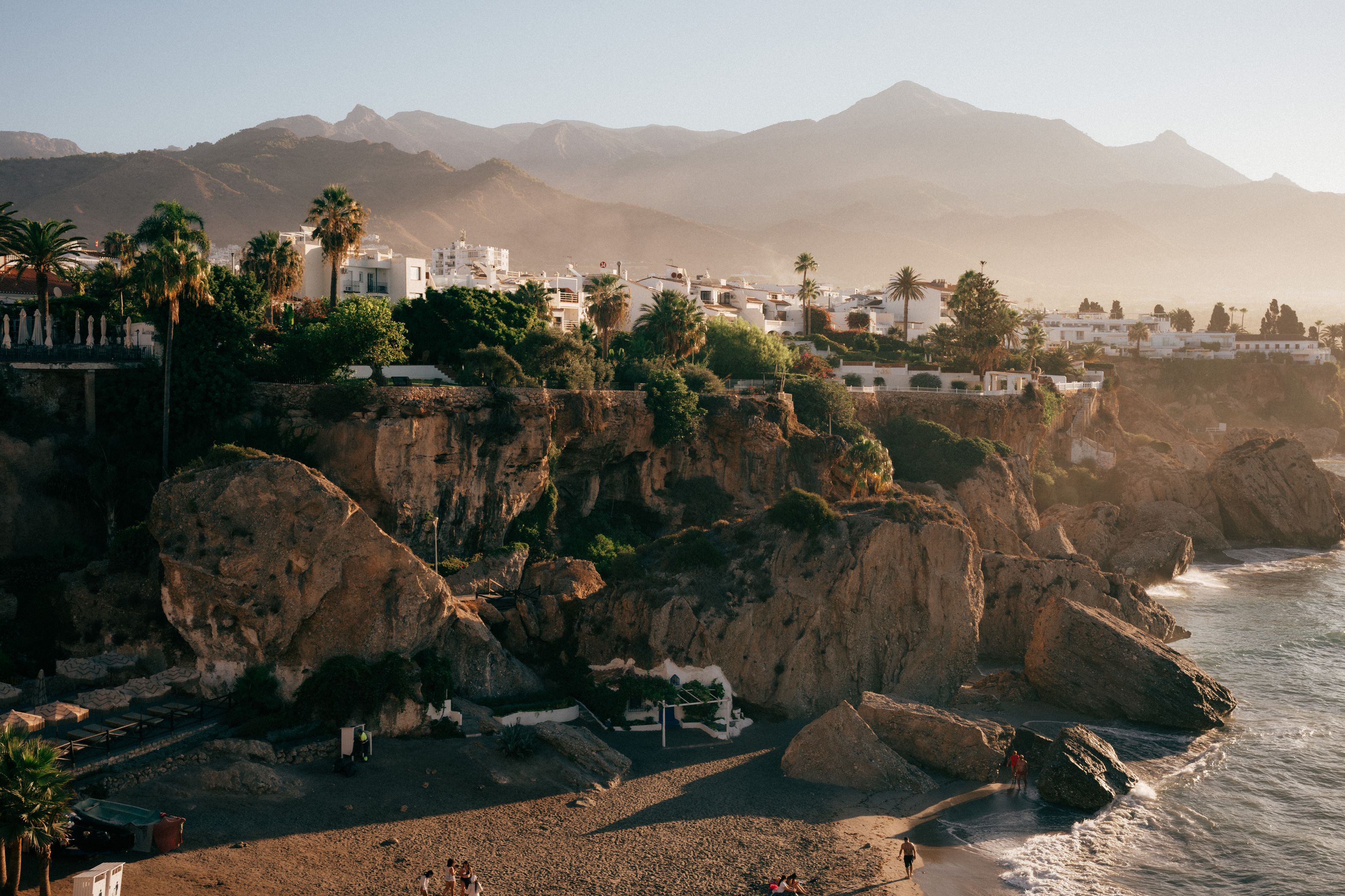 White houses and palm trees on the cliffs above a small beach at Nerja, with mountains behind in the early light